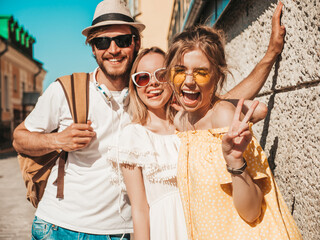 Group of young three stylish friends posing in the street. Fashion man and two cute girls dressed in casual summer clothes. Smiling models having fun in sunglasses.Cheerful women and guy outdoors