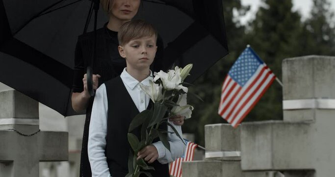 Mom and child standing near gravestone with american flag under umbrella. Widow and young boy with white lily flowers honoring husband and father at cemetery. Concept of memorial day