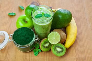Green smoothie in a glass surrounded by fruit and spirulina in a jar