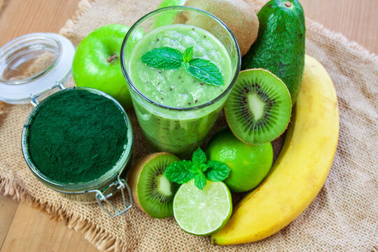 Green Smoothie In A Glass Surrounded By Fruit And Spirulina In A Jar