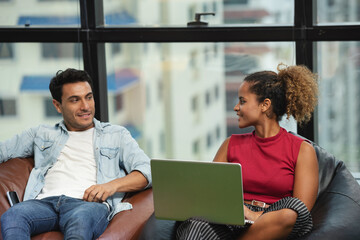 Couple of young designers sitting sofa working at modern office, two coworkers discussing fun project over a laptop and smartphone, little team of businesspeople smiling together successful meeting.
