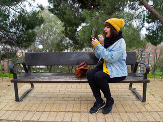 Young adult woman with yellow woolen cap sitting on a park bench painting her lips. Autumn urban concept.