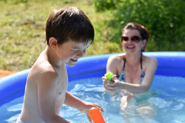 Little boy and his mom play with a water gun in pool. Cheerful child play with mom in a little pool in garden