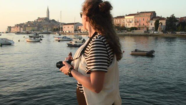 Young woman taking a picture of Rovinj marina and city view with boats and St. Euphemia cathedral at sunset. Istria, Croatia. Tracking shot with gimbal