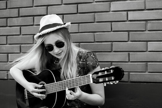 Beautiful teenage blonde long haired girl playing acoustic guitar outdoor over blue wooden fence. Youth concept. Black and white image.