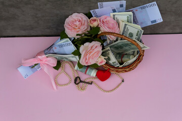 View from above. There is a basket with money on the table, a bouquet of pink roses wrapped in banknotes, a red heart, beads and a key..