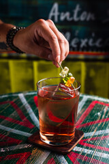 A transparent cocktail in a highball glass with a big ice cube. A hand putting gummy bears garnish on top of a drink