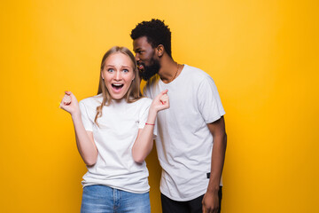 Cheerful young mixed couple whispering secret behind her hand sharing news posing isolated on yellow background. People lifestyle concept. - obrazy, fototapety, plakaty