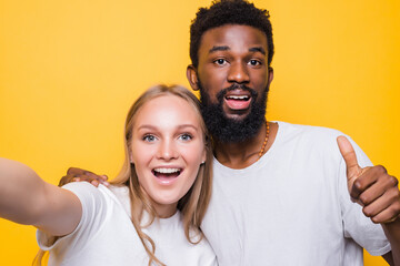 Selfie Fun. Cheerful interrational couple taking self-portrait together, looking at camera and smiling, posing over yellow background