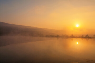 Sunrise over foggy lake. Canes on foreground on the lake coast. Sun is rising up over the trees on the further river bank.