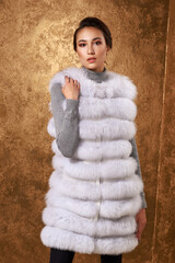 Fashion studio portrait of beautiful lady with elegant hairstyle in white luxury expensive fur coat. Winter beauty. Lady posing against golden background