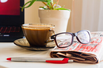 A laptop labeled news in red stands on a white table. Glasses on a black laptop. A Cup of hot coffee. Newspaper, pen.