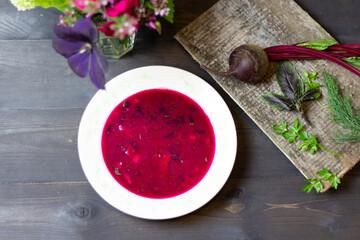 Cold red beetroot soup in white plate on black wooden background with ingredients on board and flowers