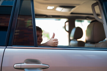 The boy sits in the back seat of a silver car and shows a thumb. Preschooler is ready for an exciting trip.