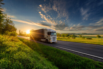 White truck driving on the asphalt road in rural landscape in the rays of the sunset