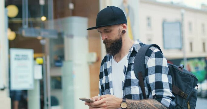 Young Caucasian handsome man with beard, tattoos and piercing tapping and scrolling on mobile phone while walking outdoor. Stylish guy hipster texting message on smartphone and chatting at street.