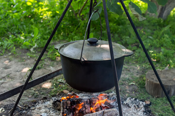 A black boiler covered with a lid on a tripod over a bonfire against