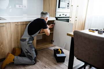 Full length shot of aged repairman in uniform working, fixing kitchen cabinet using screwdriver....