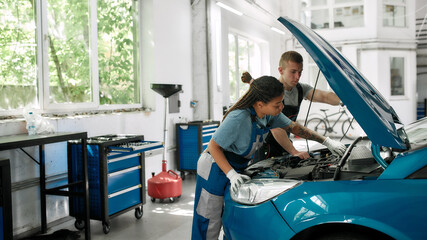 First class performance. African american woman, professional female mechanic repairing car engine, tighten, screw with spanner while her colleague holding torch under car hood at service station