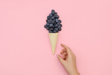 A hand reaches for a ice cream cone with blueberries, on a pink background.
