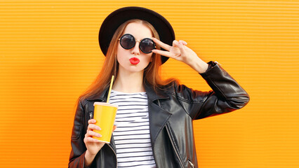 Stylish portrait of young woman with cup of juice wearing a black round hat, sunglasses over orange wall background