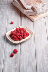 Raspberry fruits in plate near to old cutting boards, healthy pile of summer berries on grey wooden background, angle view