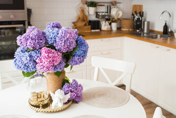 Beautiful pink, blue, purple bouquet of hydrangeas on a gold tray on the table. In the background-the interior of a modern white kitchen in the Scandinavian style. The concept of home comfort.