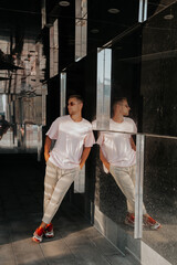 Trendy young male in casual outfit with sunglasses leaning on glass wall of modern building at daytime.