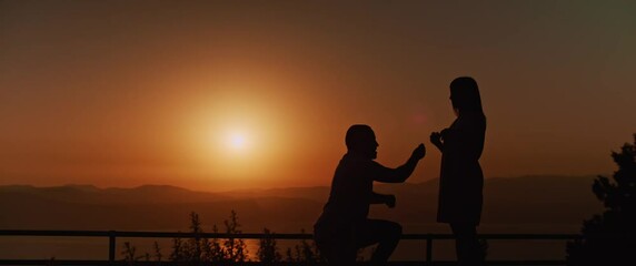 Silhouette of a man proposing to his girlfriend, putting ring on girl's finger, they get engaged at sunset, with beautiful mountains and a lake in the background. Slow motion. 