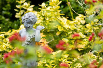 Decorative classic angel garden sculpture with out of focus red flowers in the foreground and greenery plants in the background