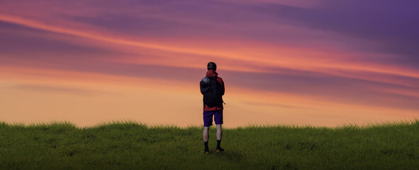 Hiker with Backpack standing on green grass and sunset.