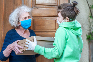 Teen grandson in mask gives grandmother food boxes of pizzas through the doorway of a house. Volunteer helps single elderly people. Family support, caring. Quarantined, isolated. Coronavirus covid-19