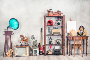 Antique media devices, writers tools, gramophone, film projector, old Teddy Bear toys and white canvas blank on easel, violin and guitar front concrete wall background. Vintage style filtered photo - obrazy, fototapety, plakaty