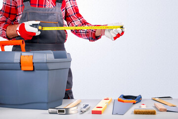 Repairman holding tape measure and toolbox against grey wall.