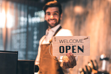 Business owner attractive young Asian man in apron hanging we're open sign on front door welcoming clients to new cafe. Happy waiter with protective face mask holding open sign while stand at cafe .