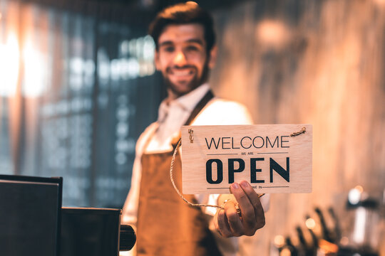 Business owner attractive young Asian man in apron hanging we're open sign on front door  welcoming clients to new cafe. Happy waiter with protective face mask holding open sign while stand at cafe .