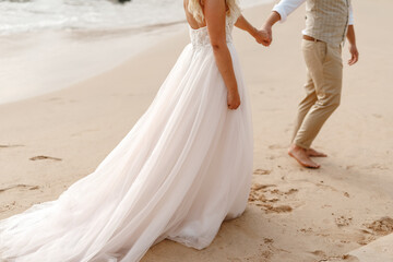 Fototapeta na wymiar Honeymoon couple holding hands walking on perfect white sand beach, cropped image, unrecognisable. Honeymoon, wedding couple on the beach