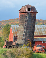 Amusing scene in rural Vermont with leaning silo, old weathered red barn, and vintage fire engine. Heavy wire wraps around silo and attaches to rusty dump truck to keep tipsy silo from falling over. © Jerrry G