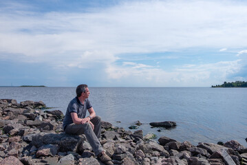 A lonely middle-aged man sits on a rocky beach on a summer day and looks thoughtfully into the distance. Loneliness, unity with nature concept. Horizontal orientation, selective focus.