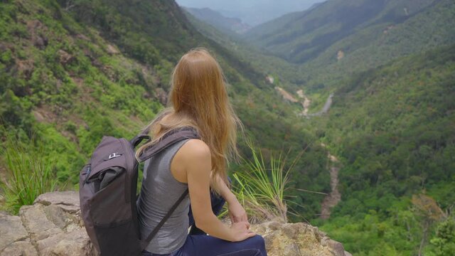 Young woman-traveler with a backpack in the mountains. She is looking at the majestic valley down there. Travel concept