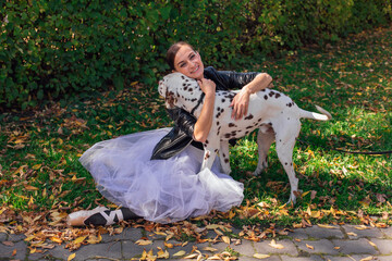 Fototapeta premium Ballerina with Dalmatian dog in the Park. Woman ballerina in a white ballet skirt and black leather jacket and in pointe shoes in autumn park hugging her spotty dalmatian dog.