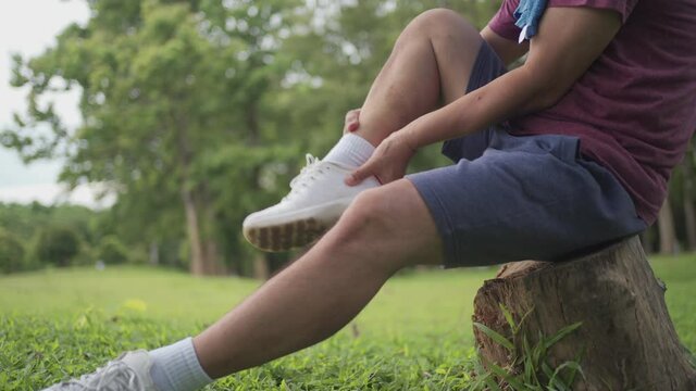 Asian man twisted his ankle during exercise Inside the park sitting on tree trunk with lawn on the background, ankle pain, joint ligament problem, painful face expression, risk of accident