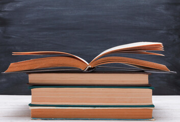 Naklejka premium Stack of books with open book on top on white wooden desk against blackboard background. Side view, copy space, close-up. Learning, education, knowledge, library, love reading concept