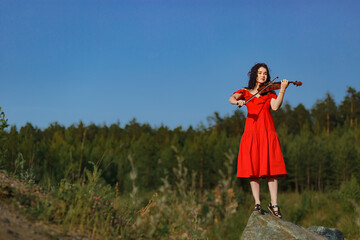 brunette girl with lush curly hair stands on the rocks near the lake in summer in a bright red dress with a violin, professional musician violinist