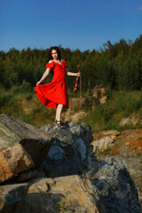 Naklejka premium brunette girl with lush curly hair stands on the rocks near the lake in summer in a bright red dress with a violin, professional musician violinist
