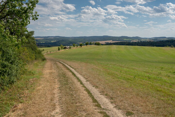 A country road in fields of central Bohemia, with wooded hills on the horizon. There are trees along the road and a pile of logs in the distance. Shot on a summer day, with a partially cloudy sky.