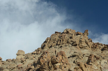 A rocky mountain slope is seen against a blue sky. A barely visible string of prayer flags tied to the rocks show the size of the boulders. The sky is blue, with wispy white clouds.