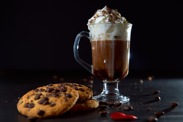 Close up of a glass cup of coffee with whipped cream and chocolate on it, chocolate chip cookies and roasted coffee beans on dark background. Concept of ready to eat food, tasty snack. Selective focus