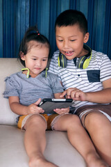 Asian children with headphones using tablet on sofa in countryside home, older brother telling his little sister stories on tablet screen and teaching how to play video.