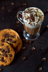Close up of a glass cup of coffee with whipped cream and chocolate on it, chocolate chip cookies and roasted coffee beans on dark background. Concept of ready to eat food, tasty snack. Selective focus